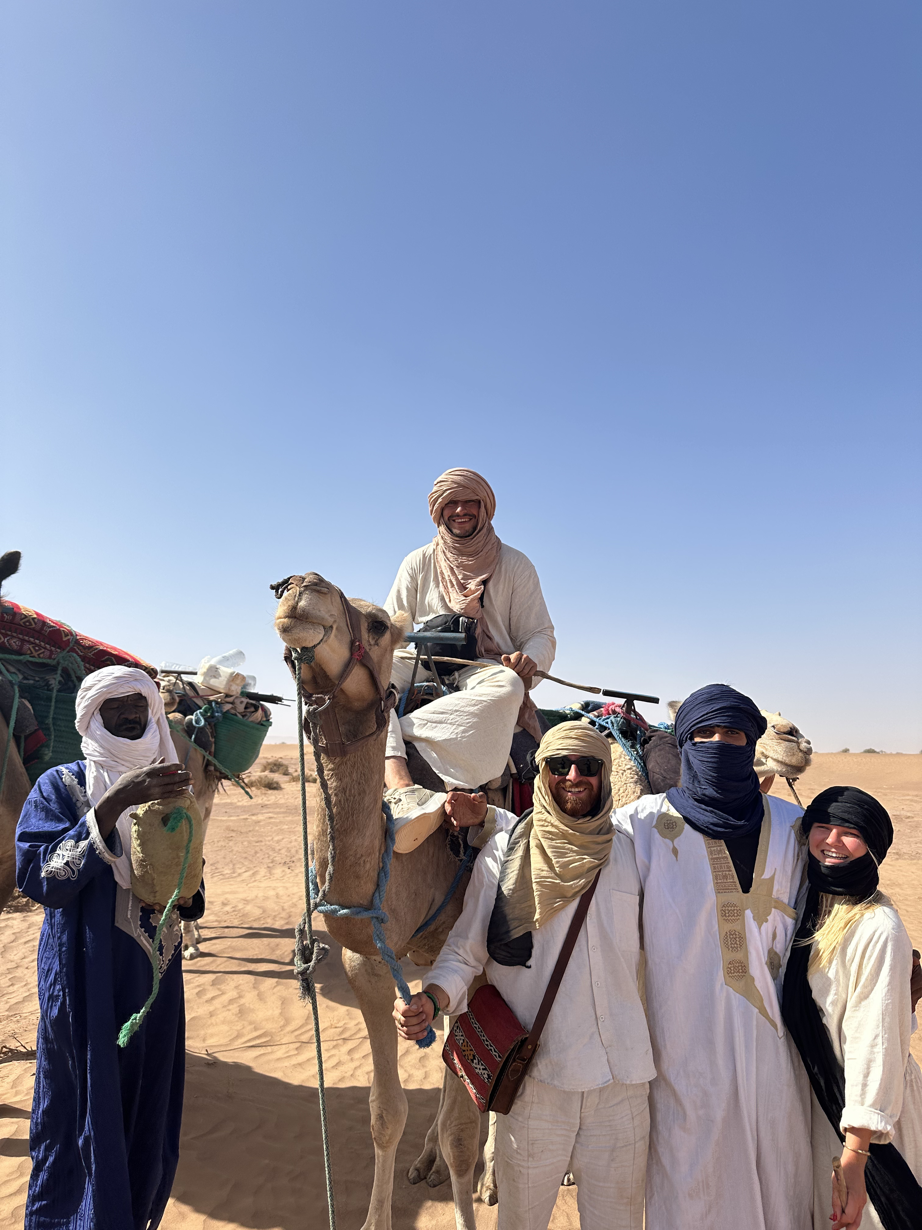 Travelers and guides gathered beside a camel caravan beneath a wide blue sky.