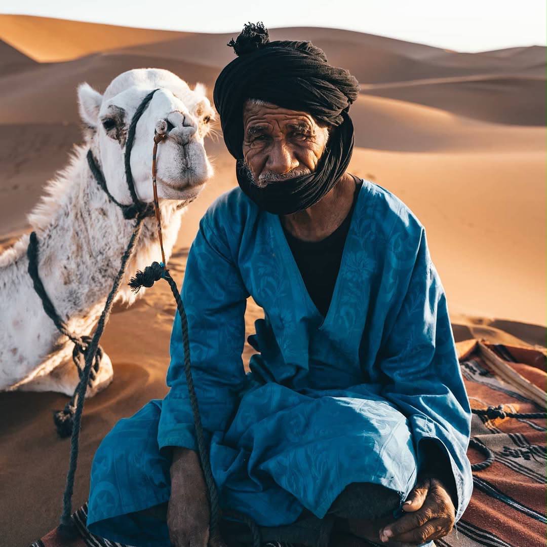 Mokhtar, senior nomad guide, seated in the Sahara beside a camel.